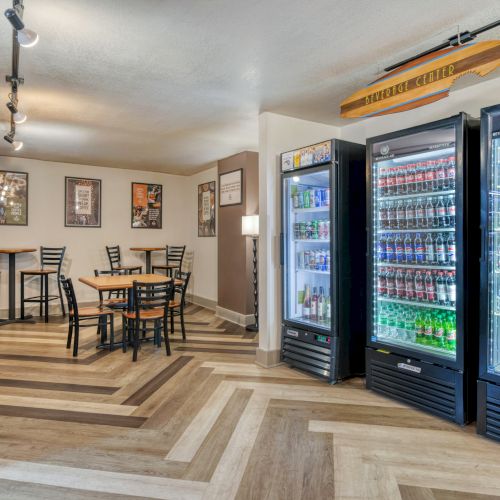 The image shows a room with tables, chairs, and stocked refrigerators. Wall art and track lighting are visible in a clean, modern setting.