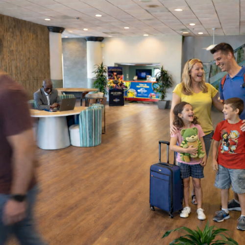 A family with two kids and luggage stands in a hotel lobby, smiling. An unrecognizable man walks by, while another sits at a desk.