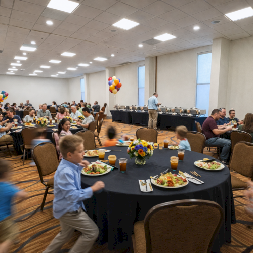 People are gathered in a banquet hall with decorated tables and balloons. Children are moving around, and food is served on tables.