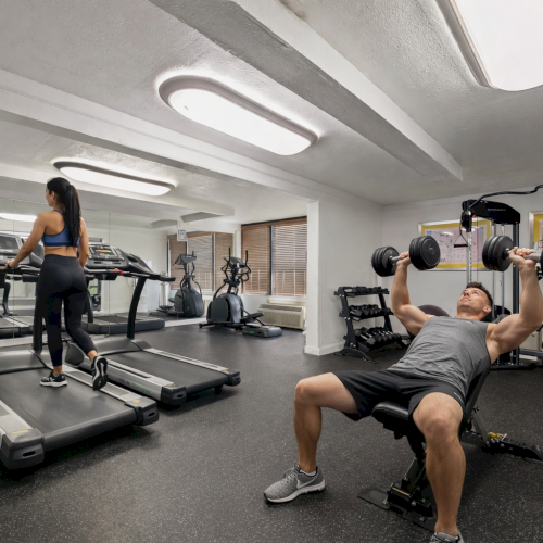 A woman walks on a treadmill and a man lifts dumbbells on a bench in a bright gym with mirrors and various exercise equipment.